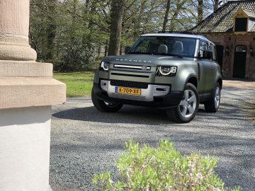 Een Land Rover Defender 90 met kenteken K-709-XF staat geparkeerd op een grindoprit buiten een gebouw met een bakstenen gevel, omgeven door bomen.