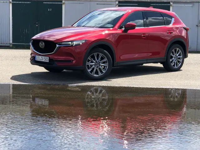 Een rode Mazda CX-5 SUV staat op de stoep geparkeerd bij een rij gesloten garagedeuren, met op de voorgrond een plas die het voertuig weerspiegelt.