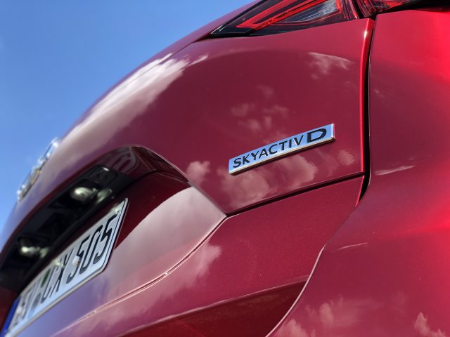 Close-up van een rode Mazda CX-5 met een 'SKYACTIV D'-badge en een deel van het achterlicht, terwijl een helderblauwe lucht wordt weerspiegeld op het gepolijste oppervlak. Het kenteken van de auto is gedeeltelijk zichtbaar.