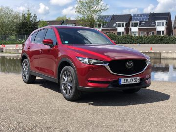 Een rode Mazda CX-5 SUV staat geparkeerd op een betonnen ondergrond nabij een reflecterende vijver met een rij huizen en bomen op de achtergrond op een zonnige dag.