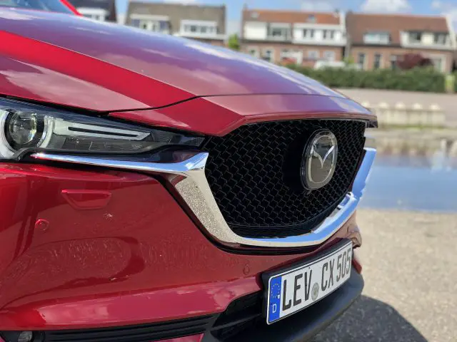 Close-up van de grille en koplamp van een rode Mazda CX-5. Op de achtergrond zijn vage woongebouwen te zien. Op het kenteken staat "LEV CX 506".