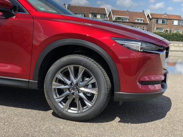 Een close-up van het linker voorwiel en de band van een rode Mazda CX-5, met woningbouw en blauwe lucht op de achtergrond.