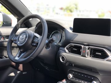 Binnenaanzicht van een Mazda CX-5 met het stuur met Mazda-logo, instrumentenpaneel, dashboard en een infotainmentscherm.