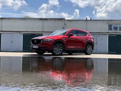         Een rode Mazda CX-5 SUV, geparkeerd voor een rij garages, werpt een levendige waterreflectie op de grond, alles onder een gedeeltelijk bewolkte hemel.