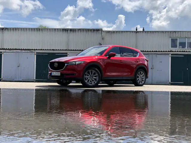 Een rode Mazda CX-5 SUV, geparkeerd voor een rij garages, werpt een levendige waterreflectie op de grond, alles onder een gedeeltelijk bewolkte hemel.