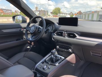 Binnenaanzicht van een Mazda CX-5 met stuur, dashboard met touchscreen, versnellingspook en stoelen. Gebouwen en een heldere hemel zijn zichtbaar door de voorruit en ramen.