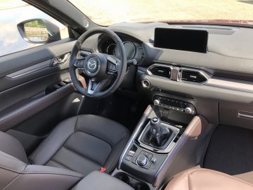 Binnenaanzicht van een Mazda CX-5 met het stuur, het dashboard, het touchscreen, de ventilatieopeningen, de versnellingspook en de middenconsole met verschillende bedieningsknoppen. De stoelen zijn bekleed met donker leer.