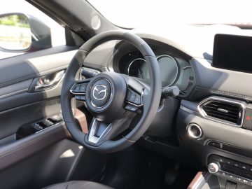 Binnenaanzicht van de Mazda CX-5 met een stuur met het Mazda-logo, dashboard, infotainmentscherm en bedieningselementen aan de bestuurderszijde.