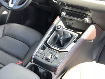 Close-up van het interieur van een Mazda CX-5 met een handmatige versnellingspook, bedieningsknoppen en bekerhouders in de middenconsole. Zonlicht verlicht de leren stoelen en de bedieningselementen op het dashboard.