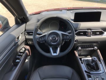 Binnenaanzicht van de Mazda CX-5 met het stuur met het Mazda-logo, het dashboard met verschillende bedieningselementen en een centraal touchscreen-display. Ook de voorstoelen en de versnellingspook zijn zichtbaar.