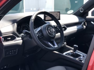 Interieur van een moderne Mazda CX-5 met een met leer bekleed stuurwiel met het Mazda-logo, dashboard, middenconsole met versnellingspook en een beeldscherm boven het dashboard.