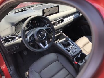 Binnenaanzicht van een Mazda CX-5, met een Mazda-stuur, een dashboard met digitale displays en een handmatige versnellingspook. De auto heeft een rood exterieur en zwart lederen stoelen.