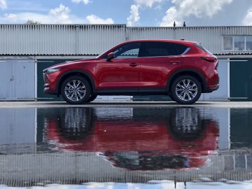 Een rode Mazda CX-5 SUV staat geparkeerd voor een gegolfd metalen hek, waarvan de weerspiegeling zichtbaar is in een grote plas op de grond eronder.