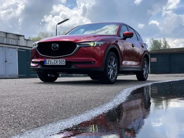 Een rode Mazda CX-5 staat geparkeerd op een nat wegdek, waarbij een grote plas de strakke lijnen van het voertuig perfect weerspiegelt.