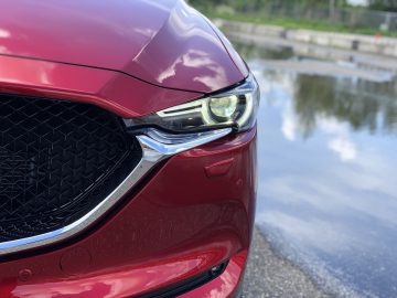 Close-up van de linkervoorkant van een rode Mazda CX-5, gericht op de koplamp en een deel van de grille, met een reflecterend wateroppervlak op de achtergrond.