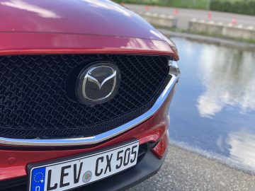 Close-up van de grille en kentekenplaat van een rode Mazda CX-5 geparkeerd nabij een watermassa.