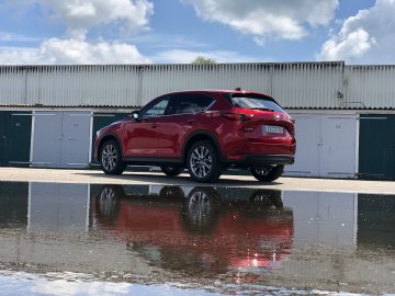 Een rode Mazda CX-5 staat geparkeerd op een verhard terrein voor witte garagedeuren, waarvan de weerspiegeling zichtbaar is in een grote plas op de grond.