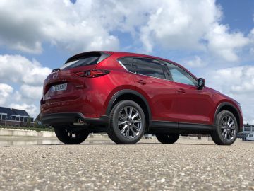 Een rode Mazda CX-5 SUV staat geparkeerd op een betonnen ondergrond onder een heldere hemel, met gebouwen op de achtergrond.