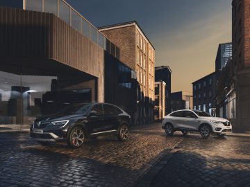 Twee auto's, waaronder een strakke Renault Arkana, staan geparkeerd in een geplaveide straat in een modern stedelijk gebied met glazen en bakstenen gebouwen onder een schemerige hemel.