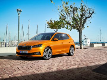 Een oranje Skoda Fabia staat geparkeerd op een klinkerbestrating bij een jachthaven met zeilboten. De achtergrond toont een helderblauwe lucht en bomen.