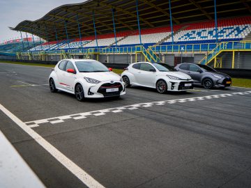 Drie auto's, waaronder een Toyota Yaris Dynamic GR Edition, staan naast elkaar op de startlijn van een racecircuit. De stands op de achtergrond zijn grotendeels leeg, met de naam “Assen” zichtbaar.