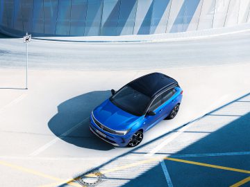 Op een lege, modern ogende parkeerplaats staat een in opvallend blauw gespoten Opel Grandland geparkeerd. Het gebied heeft een gebogen gebouw op de achtergrond en een nabijgelegen parkeerbord.