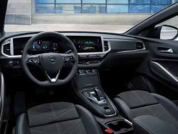 Binnenaanzicht van de Opel Grandland, met een stuur met het Opel-logo, digitaal dashboard, versnellingspook en touchscreendisplay. Zwart lederen stoelen en diverse bedieningsknoppen zijn zichtbaar.