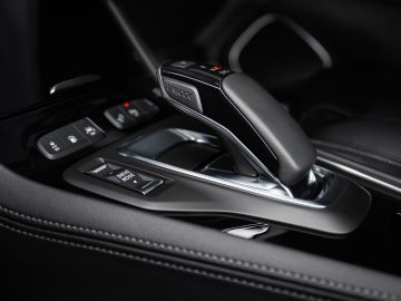 Close-up van de versnellingspook van een auto en de omliggende bedieningselementen in de Opel Grandland, met een glanzende zwarte en metallic afwerking met knoppen en stiksels zichtbaar op de binnenoppervlakken.