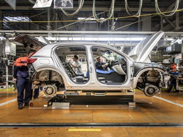Fabrieksarbeiders monteren een zilveren Volvo op een productielijn. De auto, gedeeltelijk gebouwd met zichtbare interne componenten en de motorkap en deuren open, getuigt van nauwgezet vakmanschap. Apparatuur en bovengrondse machines zijn zichtbaar, wat bijdraagt aan de precisie van het assemblageproces.