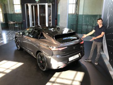 Een man staat naast een strakke, metaalgrijze DS 4 E-Tense in een showroom en gebaart naar het indrukwekkende voertuig.
