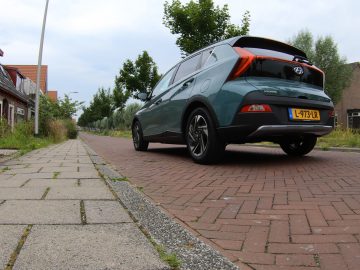 Een groene Hyundai Bayon staat geparkeerd in een geplaveide woonstraat. Het aanzicht toont de achterkant en de linkerkant van het voertuig. Gebouwen en bomen omzomen de straat.