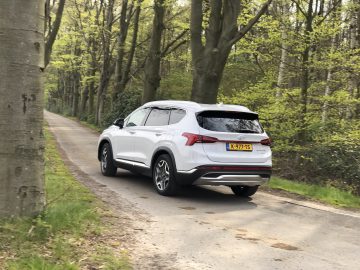 Een witte Hyundai Santa Fe Hybrid rijdt op een smalle weg tussen hoge bomen in een bosrijke omgeving. Het kenteken van het voertuig is zichtbaar.