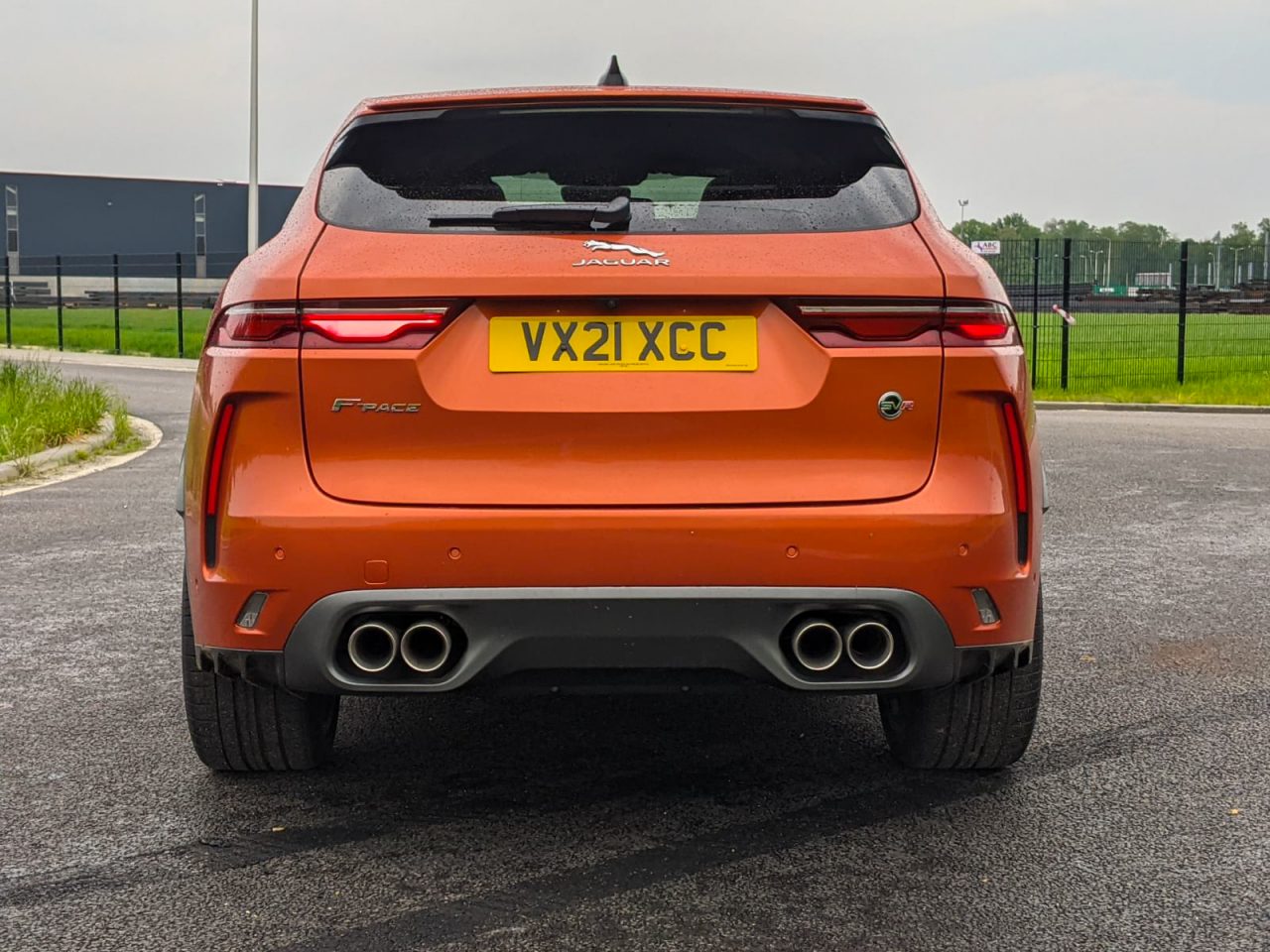 Achteraanzicht van een oranje Jaguar F-Pace met zichtbaar kenteken "VX21 XCC", geparkeerd op een asfaltoppervlak voor een omheind terrein, wat het strakke ontwerp van het voertuig laat zien.