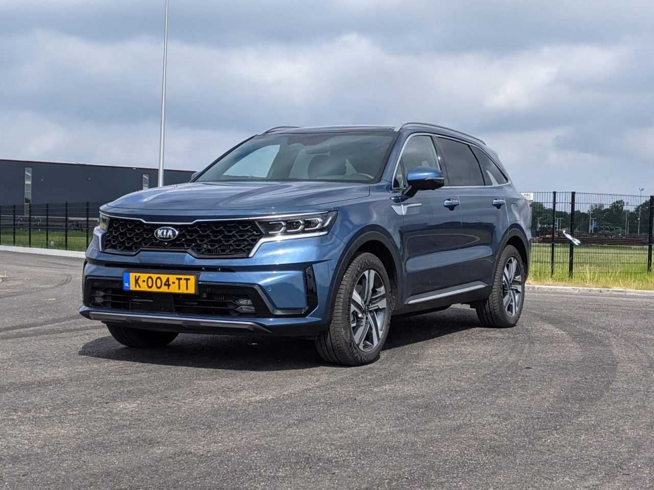 Een blauwe Kia Sorento SUV geparkeerd op een verhard terrein met een gebouw en hekwerk op de achtergrond onder een bewolkte hemel. Het voertuig heeft een geel kenteken met de tekst "K-004-TT.