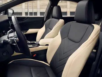 Een zicht op het interieur van een moderne Lexus NX met de voorstoelen, het stuur, het dashboard en de versnellingspook. De stoelen hebben een tweekleurige kleurstelling met beige en zwarte bekleding.