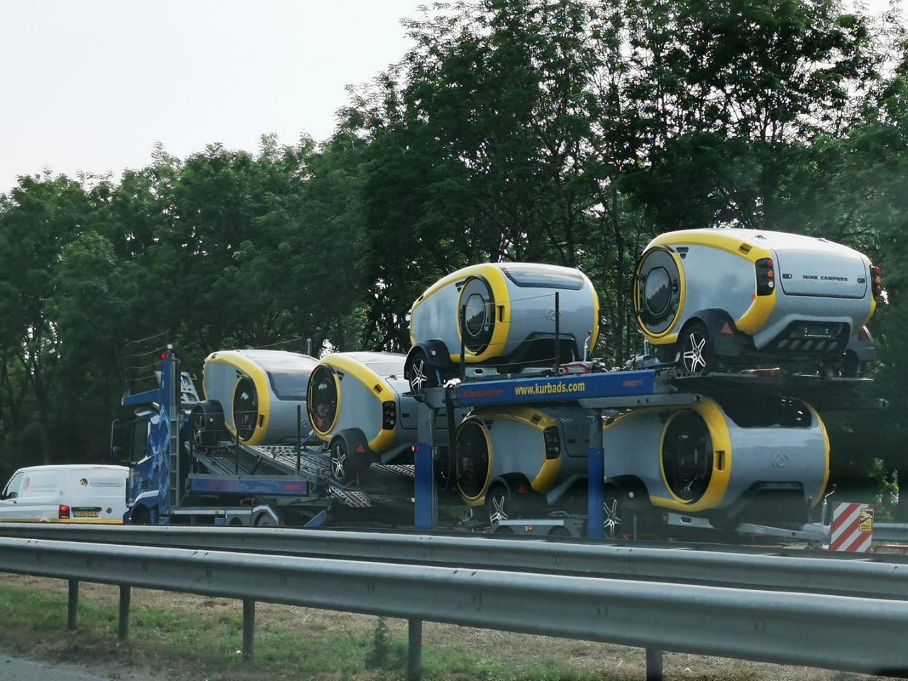 Een aanhanger vervoert zes futuristische, compacte voertuigen met gele en grijze ontwerpen op een snelweg, met zichtbare bomen op de achtergrond. Onder hen valt de gestroomlijnde Mink Sports Camper 2.0 op, die innovatie naadloos combineert met stijl.