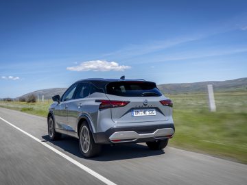 Een slanke zilveren Nissan Qashqai SUV rijdt over een landelijke weg, geflankeerd door uitgestrekte velden en een majestueuze bergketen, allemaal onder een helderblauwe lucht.