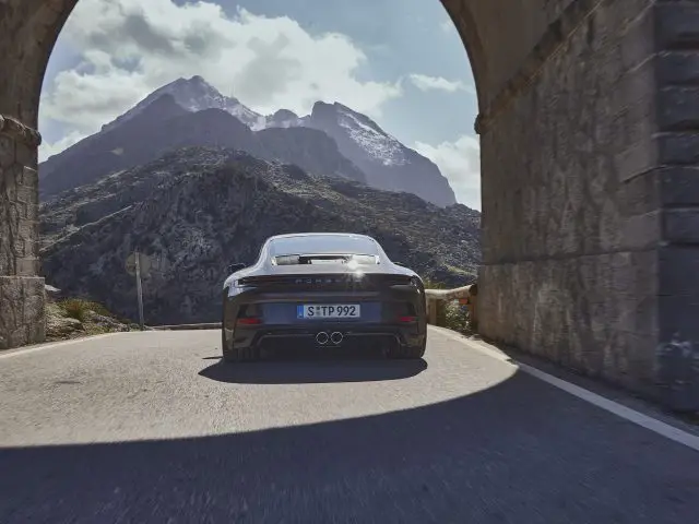 Een zwarte Porsche 911 GT3 Touring die door een stenen boog rijdt met een bergachtig landschap op de achtergrond. De weg buigt zachtjes en zonlicht verlicht het tafereel.