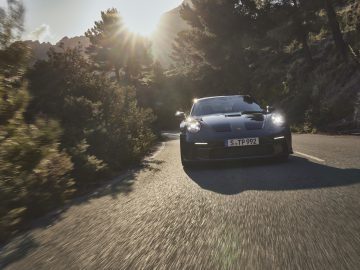 Een zwarte Porsche 911 GT3 Touring rijdt op een beboste weg in zonlicht.