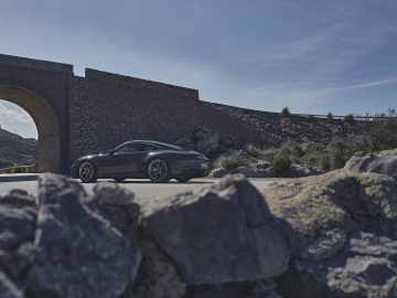 Een strakke zwarte Porsche 911 GT3 Touring staat geparkeerd op een weg bij een stenen boog onder een helderblauwe lucht, omgeven door rotsachtig terrein.