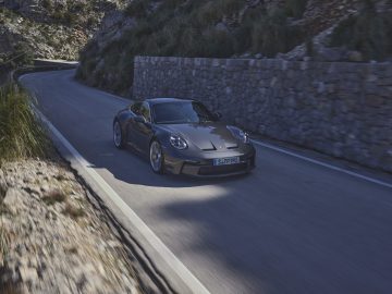 Een strakke Porsche 911 GT3 Touring rijdt over een bochtige bergweg omzoomd met stenen muren en omgeven door groen.