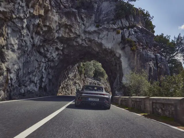 Een Porsche 911 GT3 Touring rijdt op een heldere dag door een tunnel die in een rotsachtige heuvel is uitgehouwen, met omringend groen en een blauwe lucht.