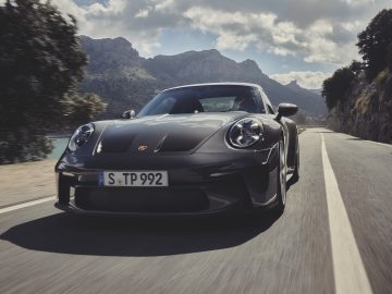 Een zwarte Porsche 911 GT3 Touring rijdt op een schilderachtige bergweg met blauwe luchten en wolken op de achtergrond.