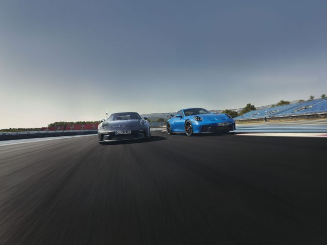 Twee auto's, een grijze en een blauwe Porsche 911 GT3 Touring, racen op een circuit onder een heldere hemel, met vage bewegingen die hoge snelheid aangeven.