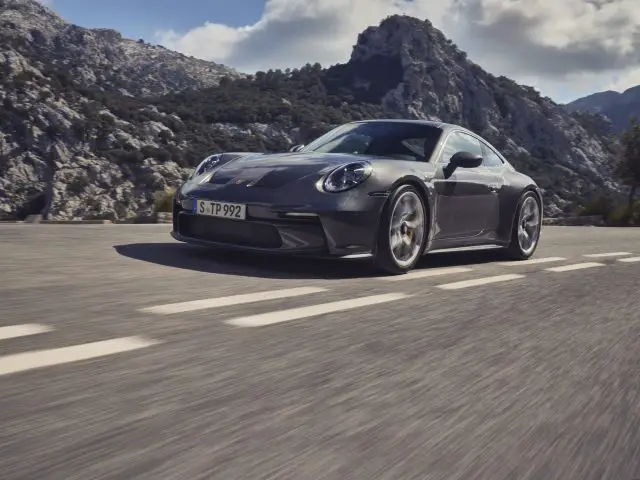 Een strakke, grijze Porsche 911 GT3 Touring rijdt over een bergweg met rotsachtige toppen en groen op de achtergrond onder een gedeeltelijk bewolkte hemel.