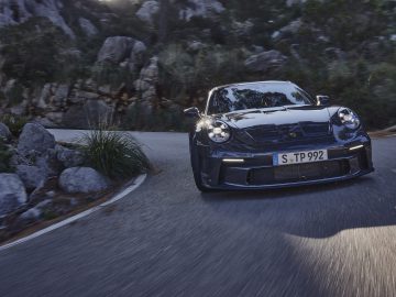 Een donkergekleurde Porsche 911 GT3 Touring navigeert over een bochtige weg door een rotsachtig, bosrijk gebied.