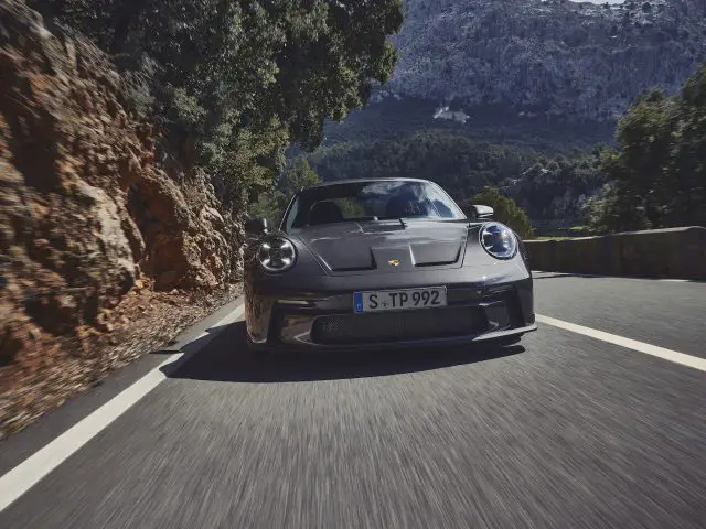 Een grijze Porsche 911 GT3 Touring rijdt op een bergweg, met bomen en rotswanden op de achtergrond.