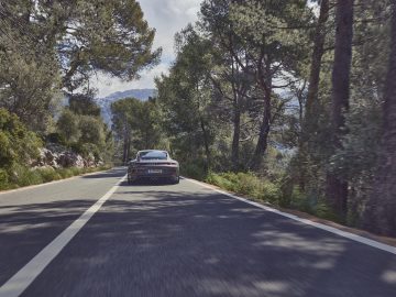 Een Porsche 911 GT3 Touring rijdt op een smalle, bochtige weg omringd door dichte bomen en bergen op de achtergrond onder een heldere hemel.