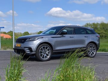 Een zilveren Range Rover Velar met getinte ramen staat geparkeerd op een verhard terrein naast hoog gras, tegen een achtergrond van een gedeeltelijk bewolkte lucht en groen.