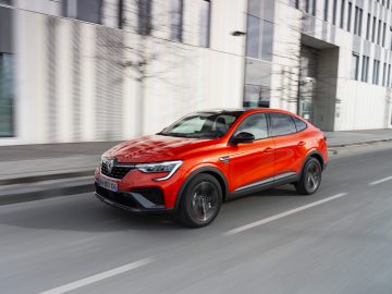 Een rode Renault Arkana rijdt door een stadsstraat vlakbij een modern gebouw met verticale raampatronen. Bomen zonder bladeren sieren het trottoir.